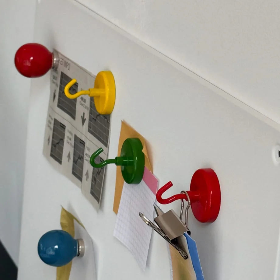 Angled close-up of red, yellow, green, and blue magnetic hooks holding papers on a whiteboard - RoomDen