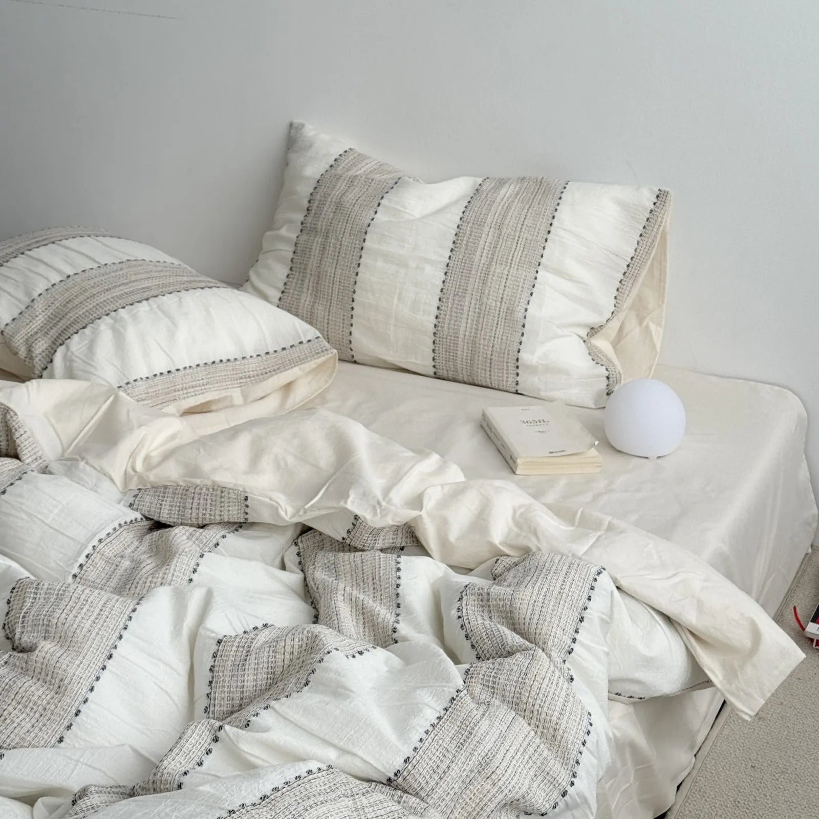 A bed dressed in an oatmeal beige textured stripe washed cotton bedding set in a bright, airy room, the Soft-Stack Method in one frame. - RoomDen