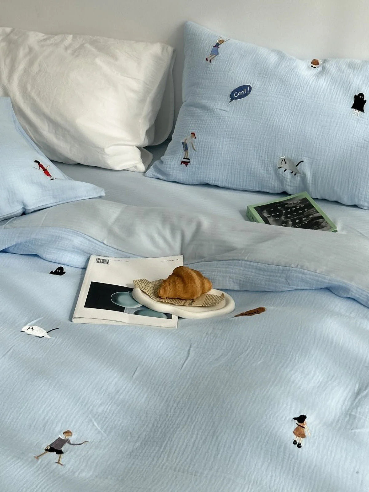 The sky blue embroidered bedding set styled with a croissant and books, creating a relaxed morning vibe.