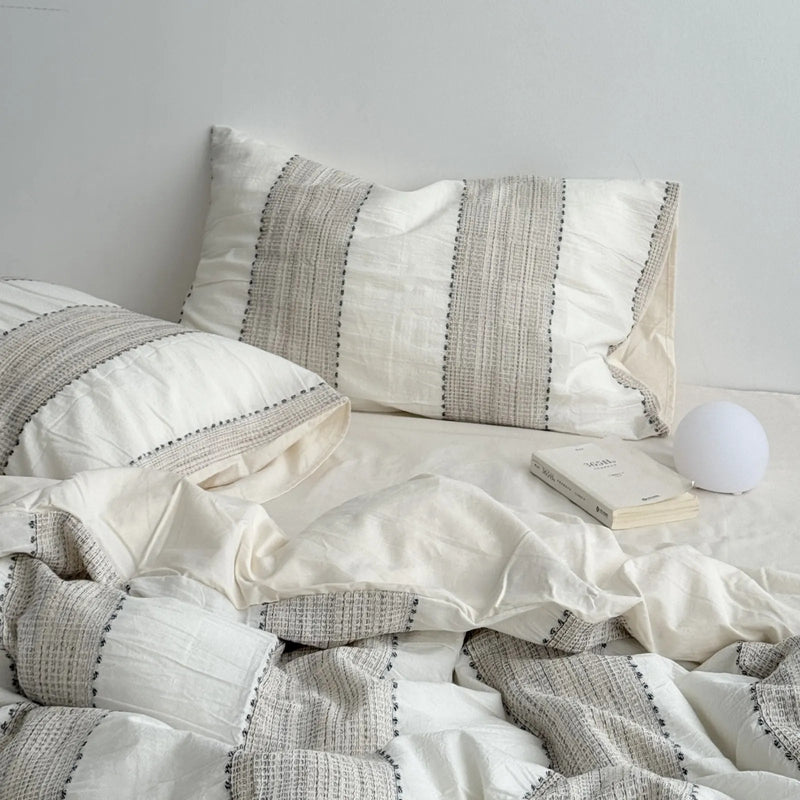 Full view of an oatmeal beige woven stripe bedding set with a small white lamp and book on the bedside. - RoomDen