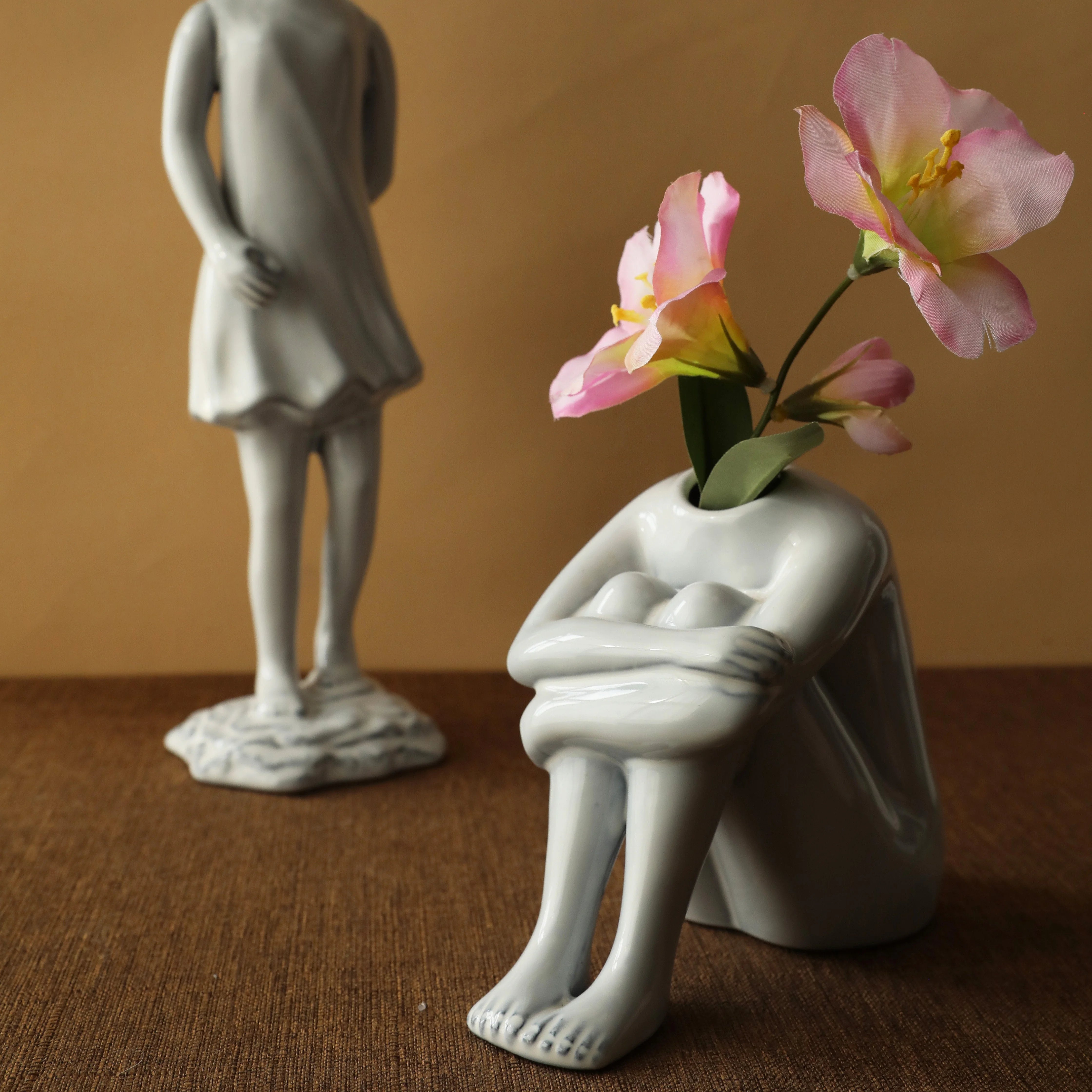 Detail shot of the crouching body form ceramic vase with pink flowers, with another vase in the background. - RoomDen