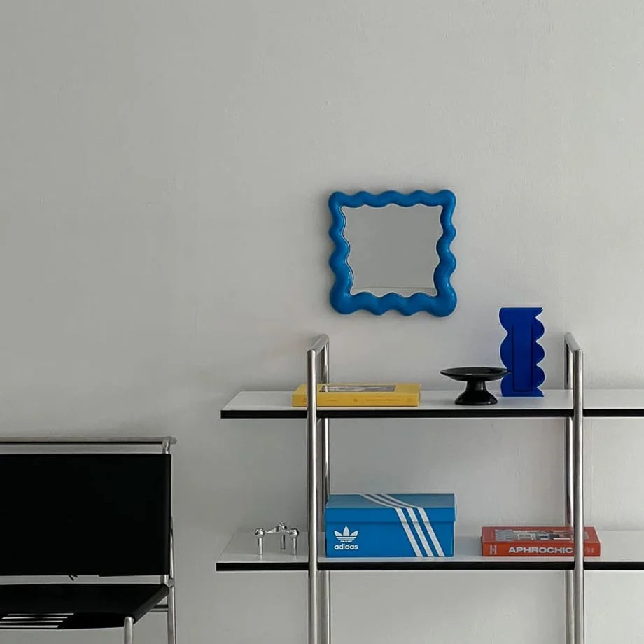 Wavy blue mirror on a white wall above a modern metal shelving unit with books and an Adidas box - RoomDen