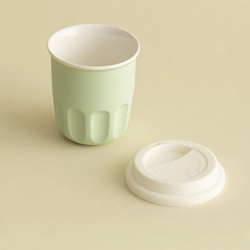 The mint green frosted ceramic cup shown with its white silicone lid placed beside it on a table. - RoomDen