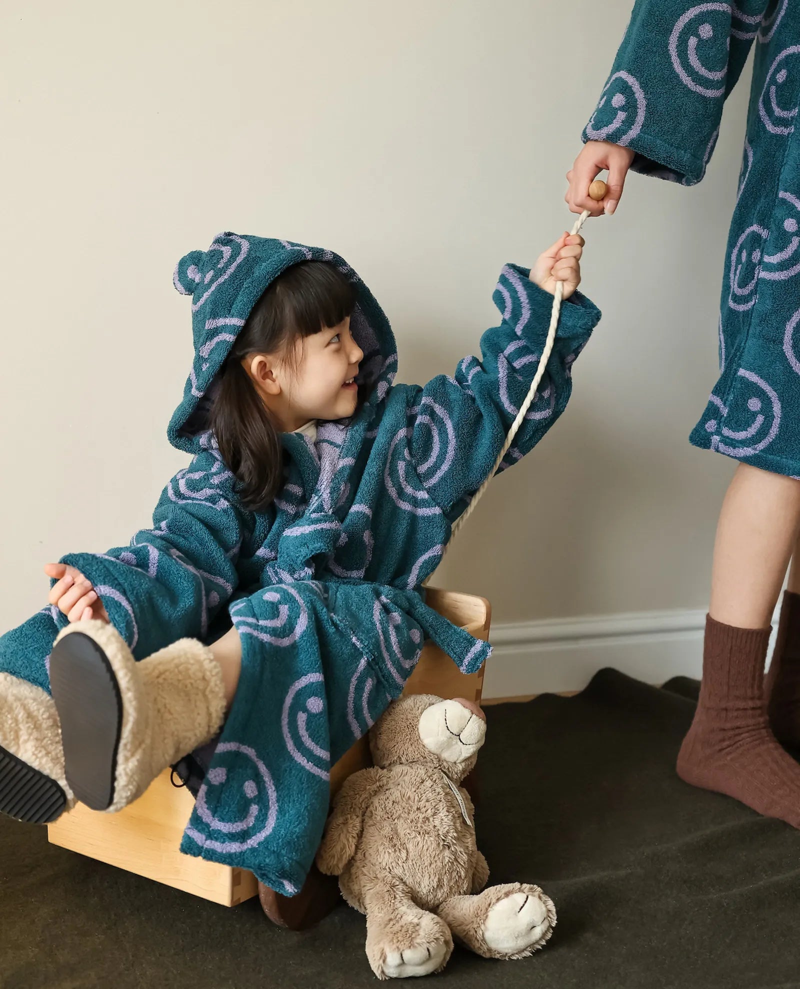 Little girl in a teal smiley face hooded robe sitting in a wooden cart being pulled by an adult in a matching robe