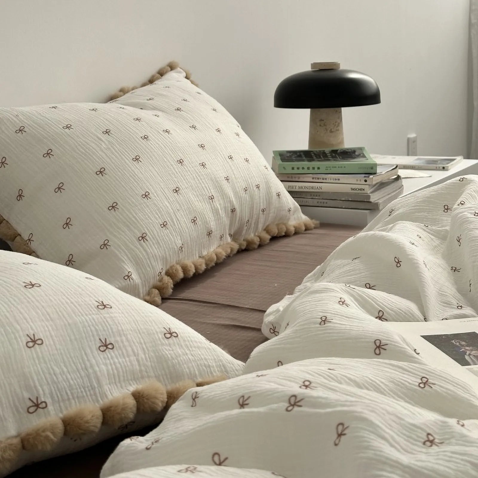 A side view of the bow print bedding focusing on a pom-pom trimmed pillow next to a bedside lamp and a stack of books. - RoomDen