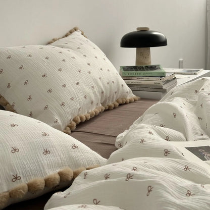 A side view of the bow print bedding focusing on a pom-pom trimmed pillow next to a bedside lamp and a stack of books. - RoomDen