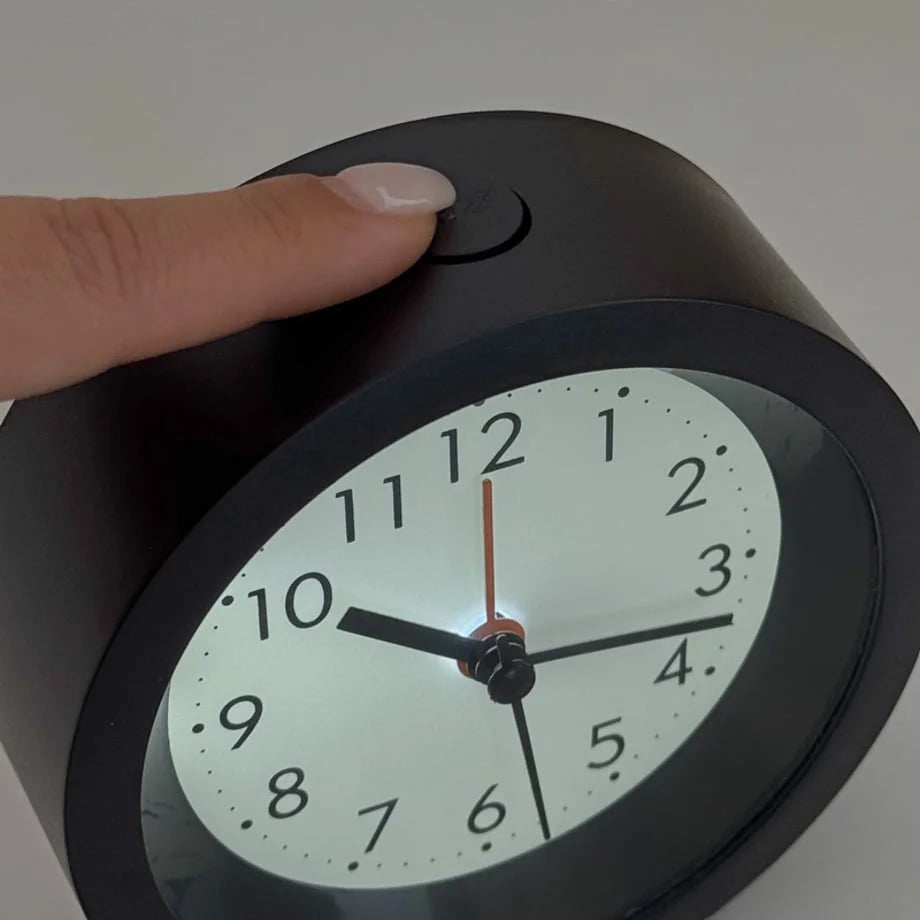 A finger pressing the top button of the black alarm clock to activate the soft night light feature. - RoomDen