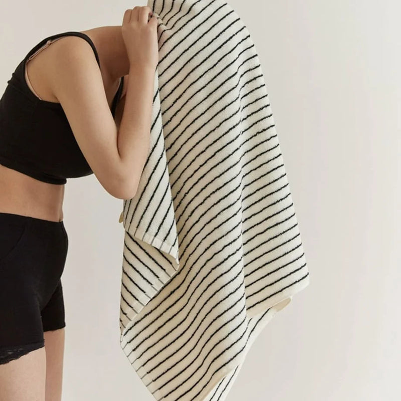 Early-morning light catching a soft, textured striped cotton bath towel as it's lifted, the room otherwise bare. — RoomDen