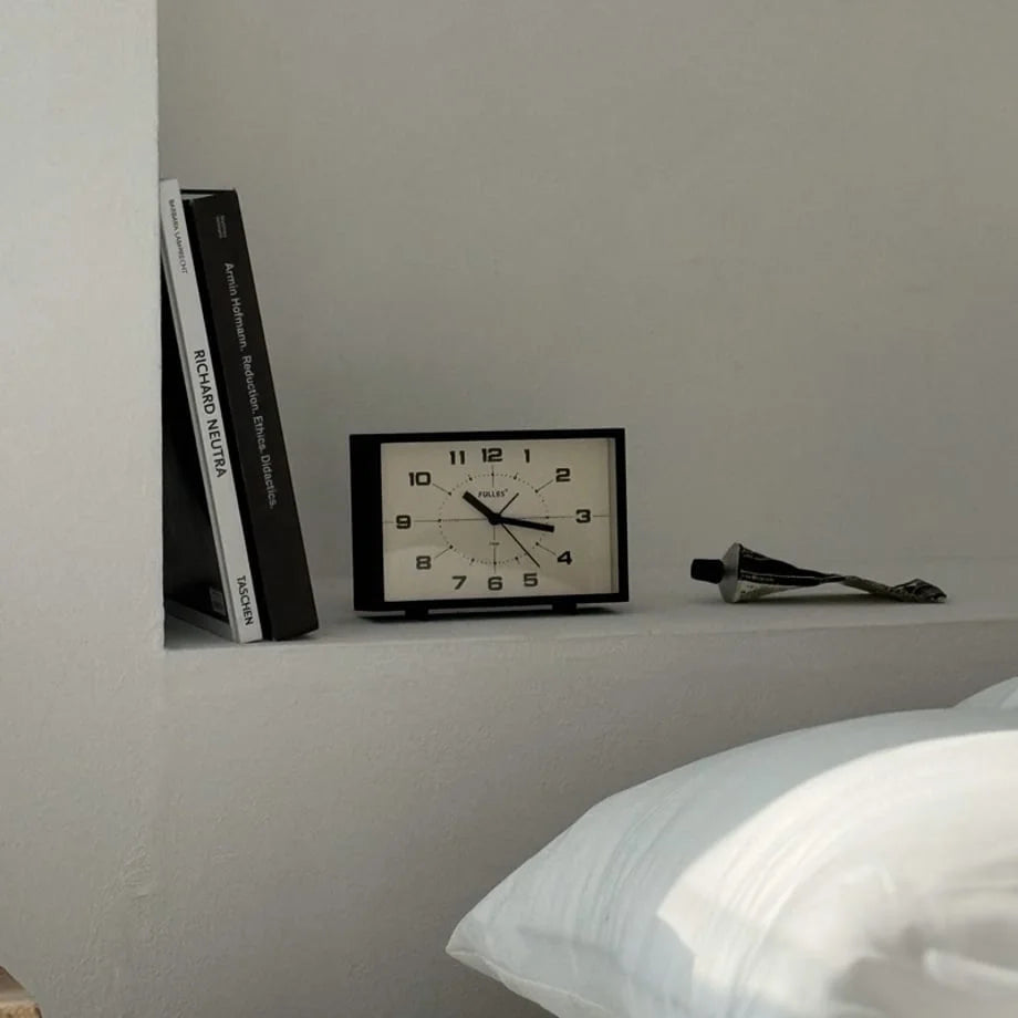 The Matte Black Retro Table Clock on a white shelf beside books and a pillow.