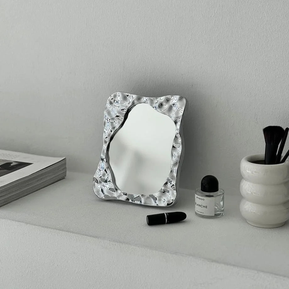 The Molten Irregular Chrome Desktop Mirror styled on a white shelf with perfume and a makeup brush holder - RoomDen