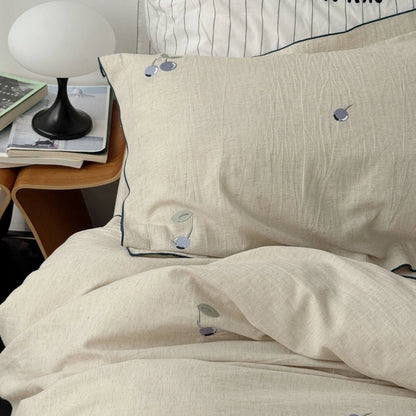 Close-up of the blueberry embroidered linen pillowcase resting on a bed, next to a modern bedside table with a lamp and books. - RoomDen