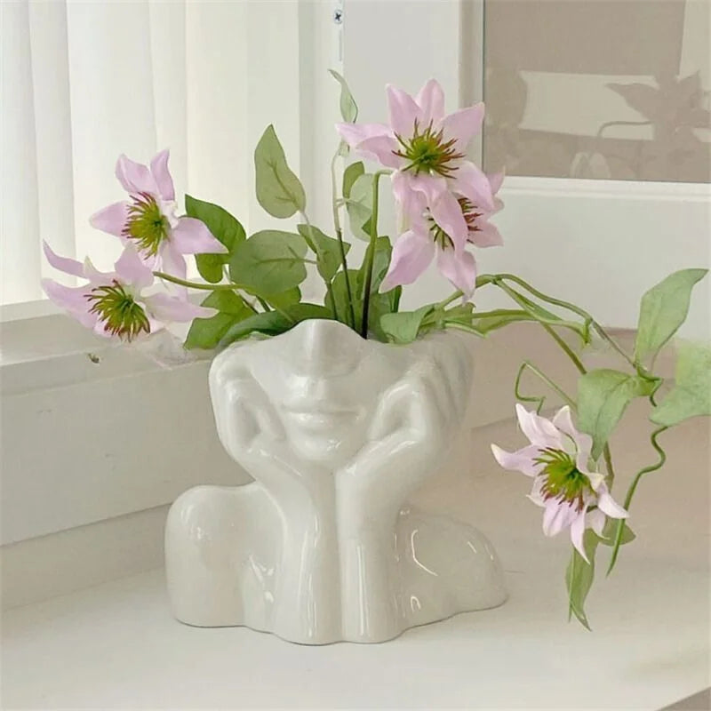 Sculptural white resin face vase holding pink flowers on a windowsill - RoomDen.
