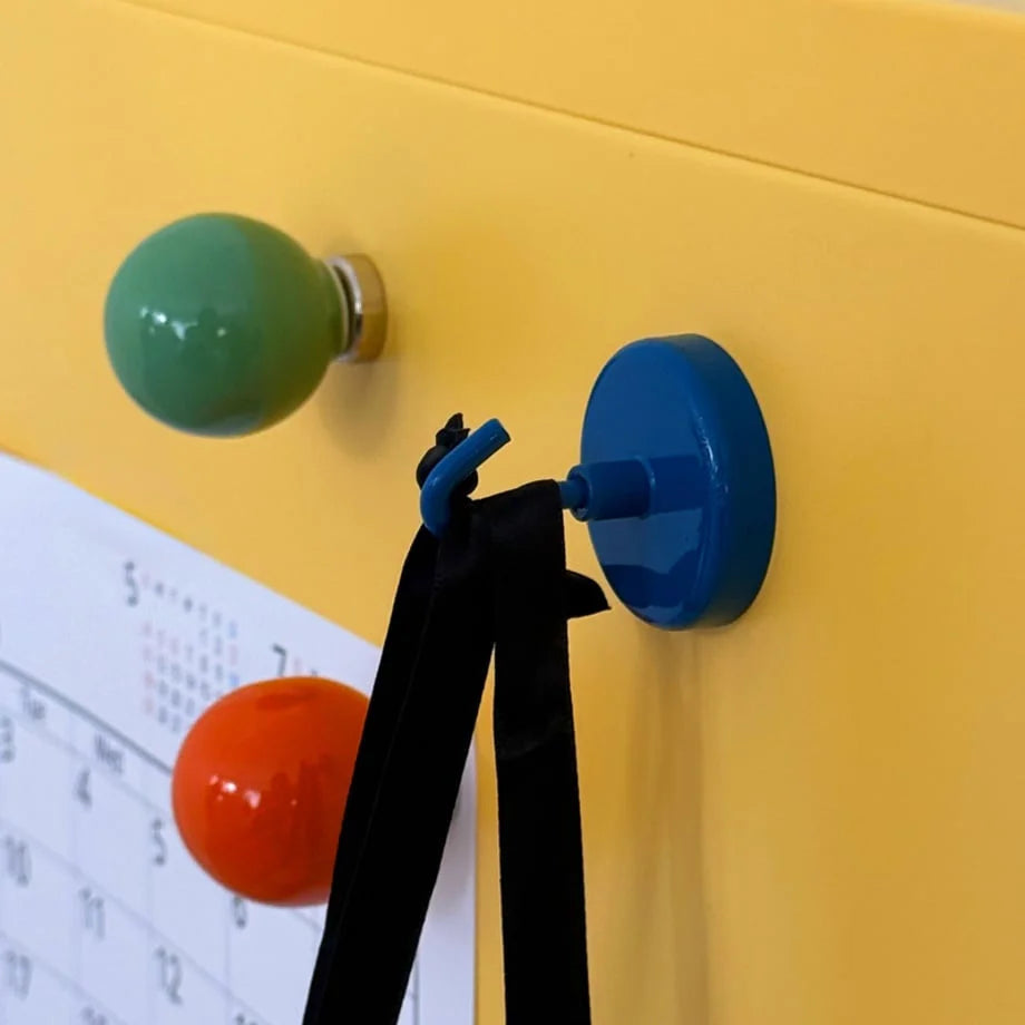Blue magnetic hook holding a black strap on a yellow metal cabinet, with green and orange magnets nearby - RoomDen