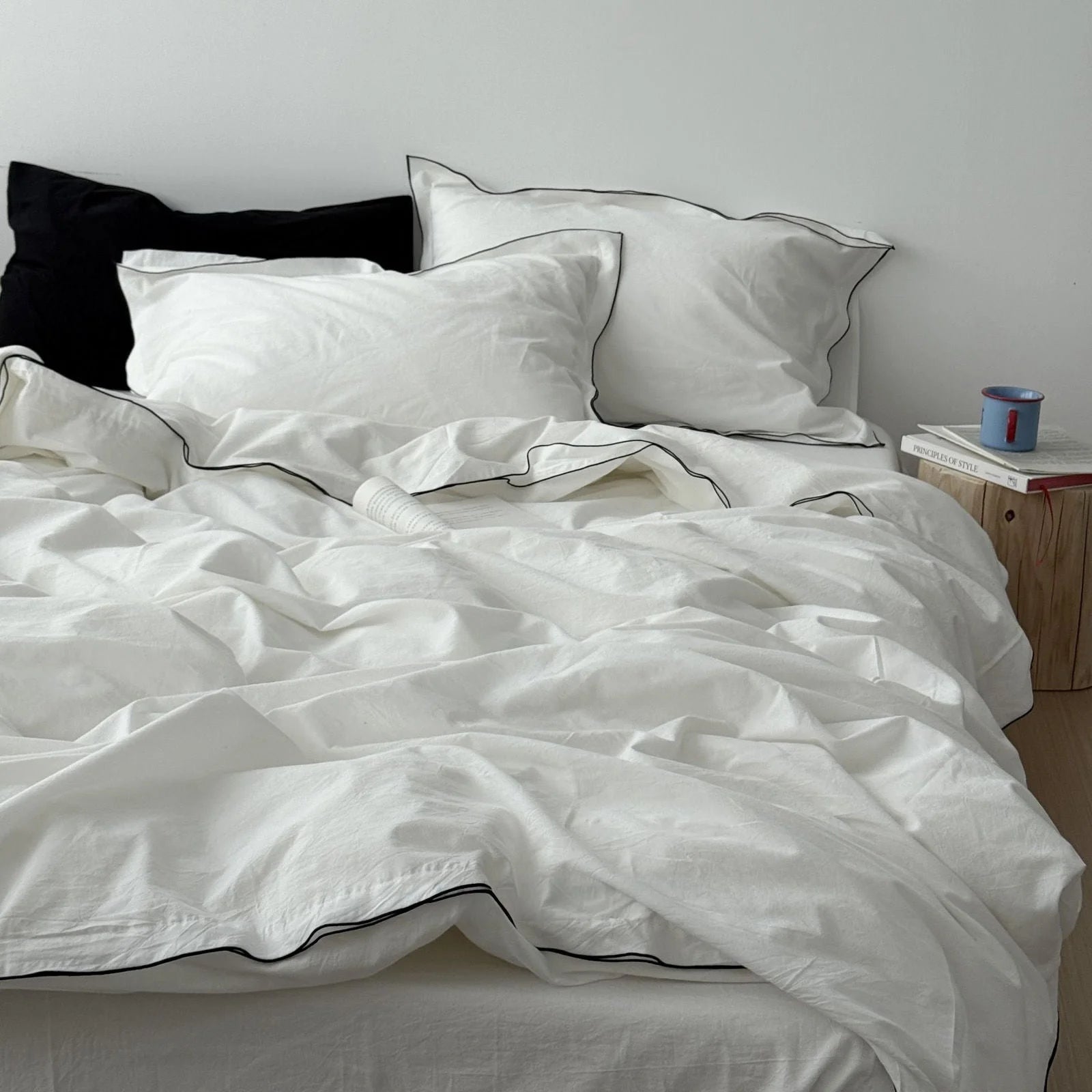Full view of a bed dressed in a white and black trim bedding set, styled with a black pillow and a wooden side table. - RoomDen