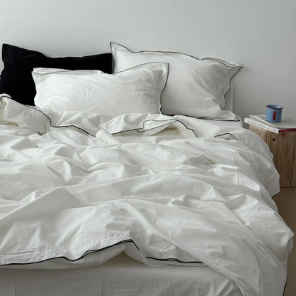 Full view of a bed dressed in a white and black trim bedding set, styled with a black pillow and a wooden side table. - RoomDen