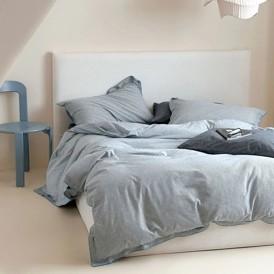 Angled shot of the Dark Grey & Blue Pinstripe Washed Cotton Bedding Set on a bed with a blue chair nearby.