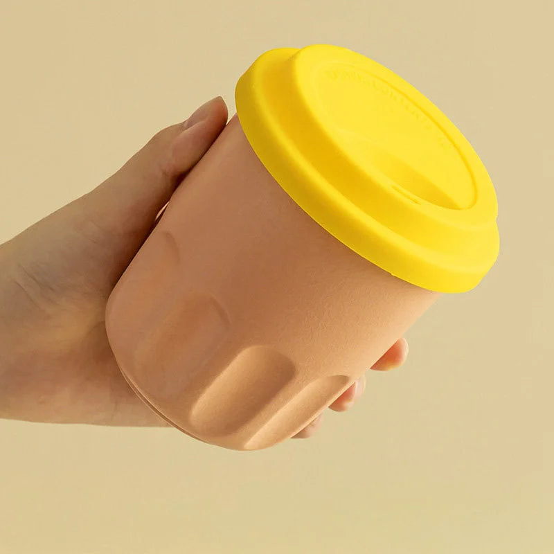 Close-up of a hand holding a peach beige frosted ceramic cup with a bright yellow lid. - RoomDen
