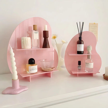 A pink wavy acrylic desktop organizer displaying perfumes and a candle on a white desk. - RoomDen