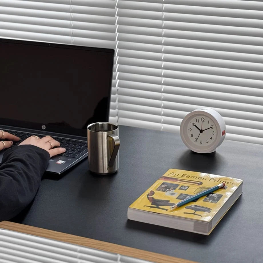 A person typing on a laptop with the white analog alarm clock keeping time on the desk. - RoomDen