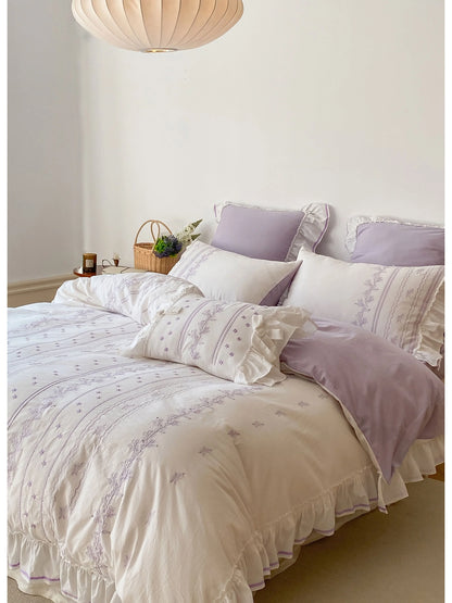 The Lavender Ruffled Floral Vine Embroidered Bedding Set on a bed next to a wicker basket with flowers.