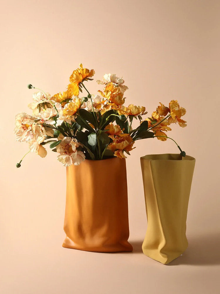Lifestyle image of the Tangerine and Mustard Yellow paper bag vases side-by-side, with the Tangerine one holding yellow and cream flowers - RoomDen.