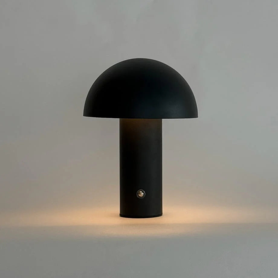 The black metal mushroom lamp turned on with a warm, ambient glow at its base against a grey background - RoomDen.