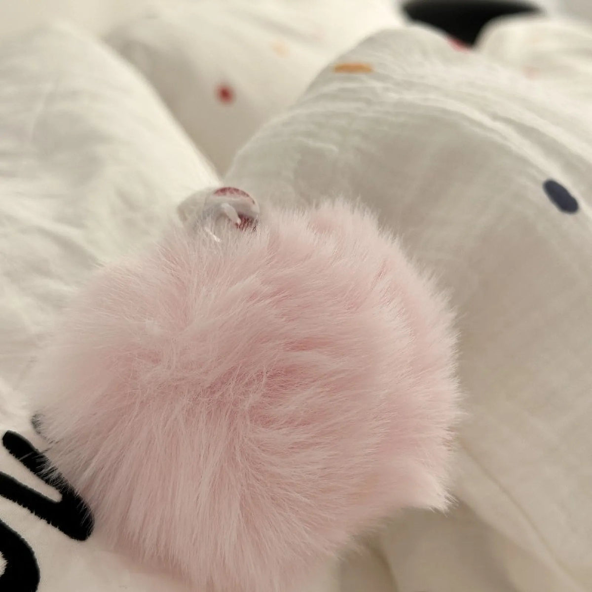Extreme close-up of a fluffy, soft pink pom-pom detail on the corner of a white pillowcase. - RoomDen