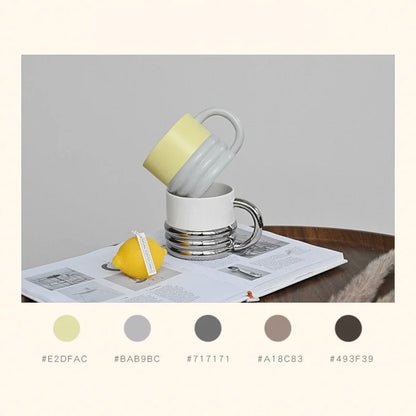 A stack of two chunky ribbed ceramic mugs, one yellow and grey, one white and chrome, on an open book - RoomDen.