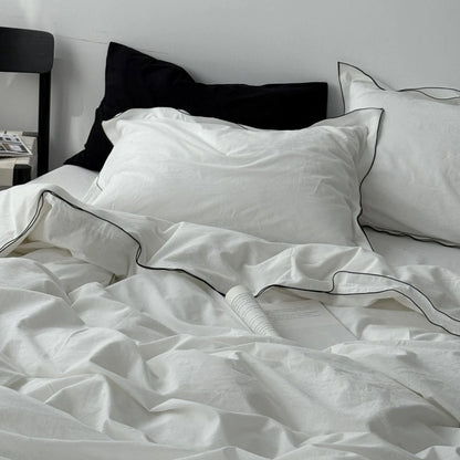 A cozy, unmade bed with a white washed cotton duvet cover and pillows, both featuring a delicate black trim. - RoomDen