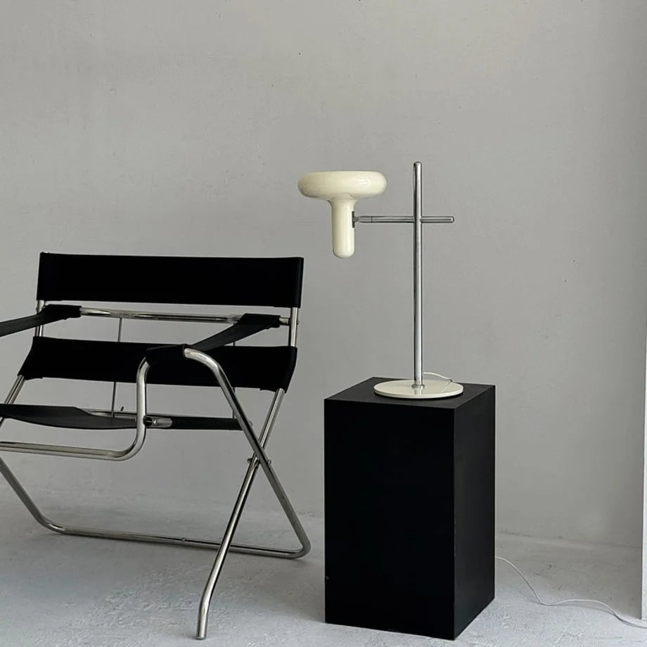 The Cream Sculptural Accent Lamp displayed on a black pedestal next to a modern chair - RoomDen.