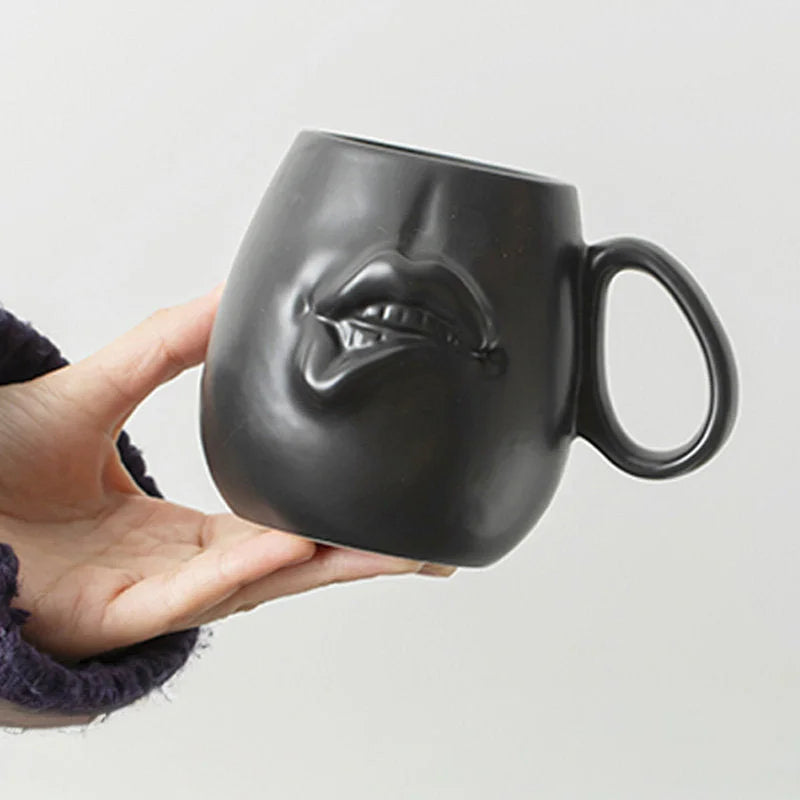 A hand holding the Matte Black Sculptural Abstract Face Ceramic Mug, showing the 3D lip detail.