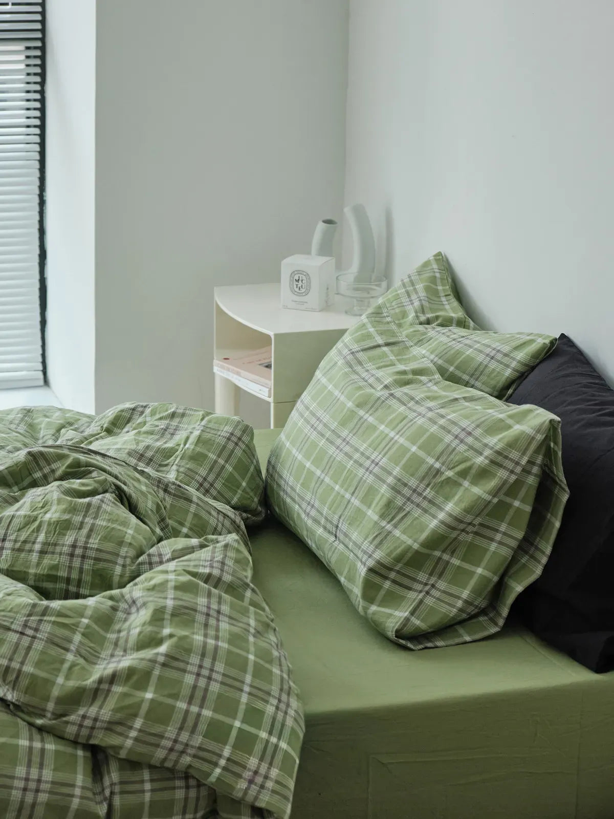 A side view of a bed made with the green plaid bedding, showing a white bedside table with a candle and vases in a bright, minimalist room. - RoomDen