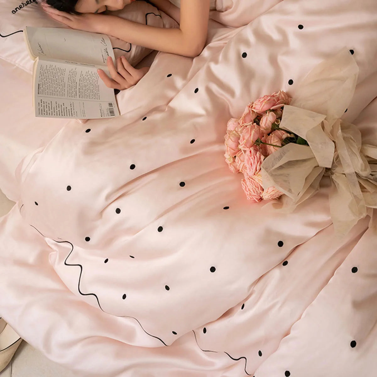A person resting on the Pastel Pink & Black silky Tencel bedding set with a book and roses. - RoomDen