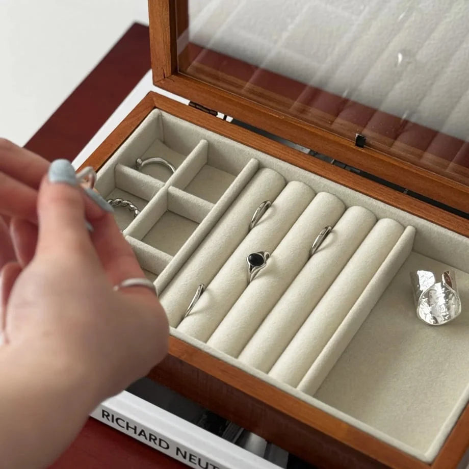 A detailed close-up of a hand selecting a ring from the soft, beige-lined compartments of the wood jewelry box.