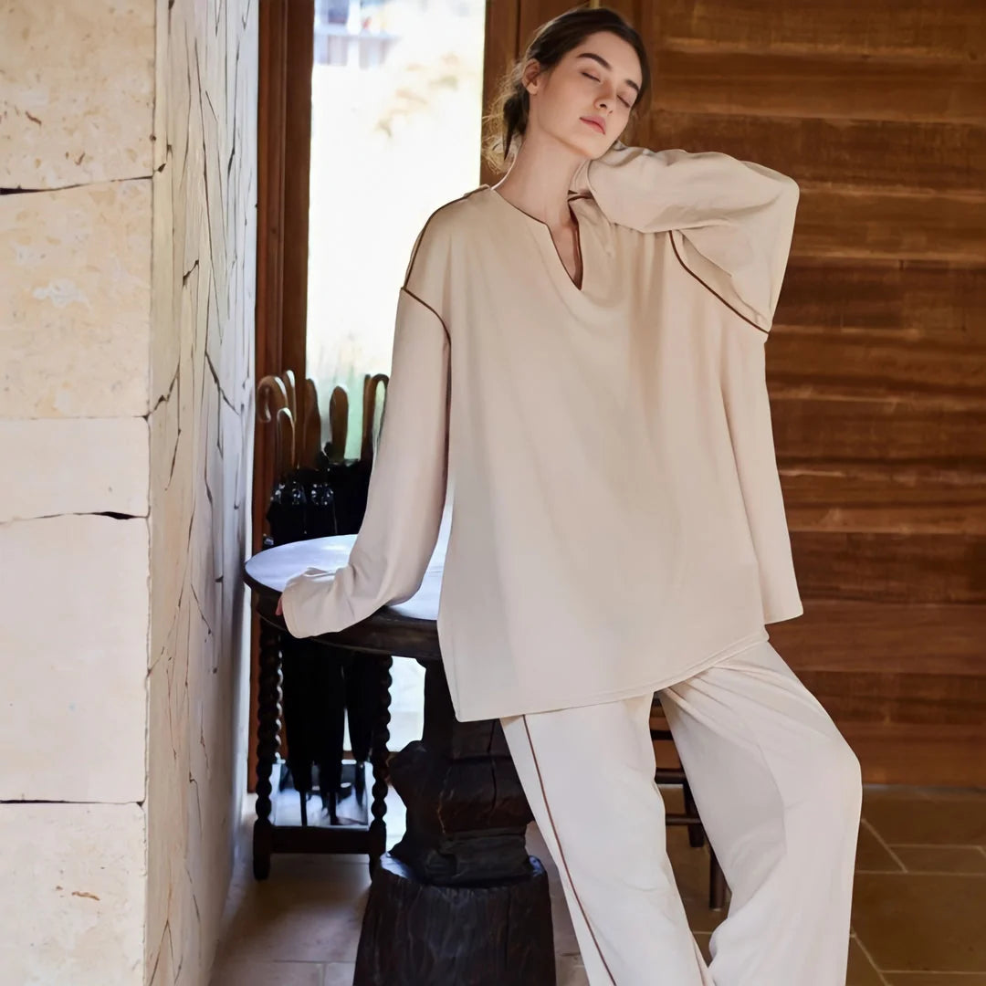 Model relaxing in the soft piped pajama set, leaning against an ornate wooden table. - RoomDen