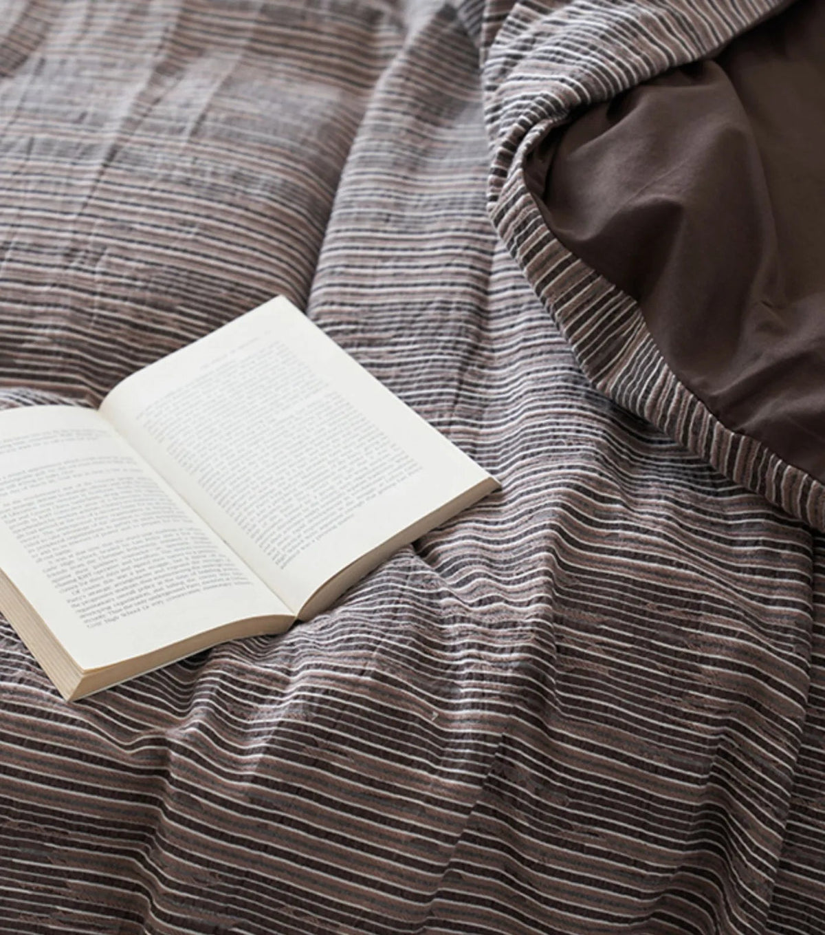 Close-up on the texture of the Mocha Brown pinstripe bedding with an open book resting on it - RoomDen