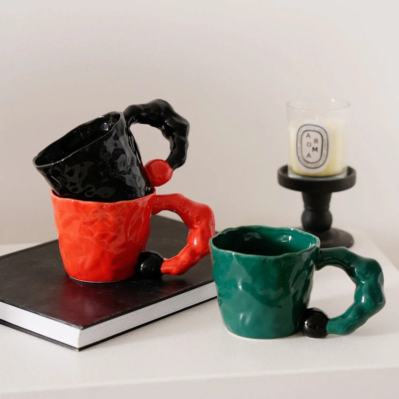 Three sculptural ceramic mugs in black, red, and green stacked on a book next to a candle - RoomDen.