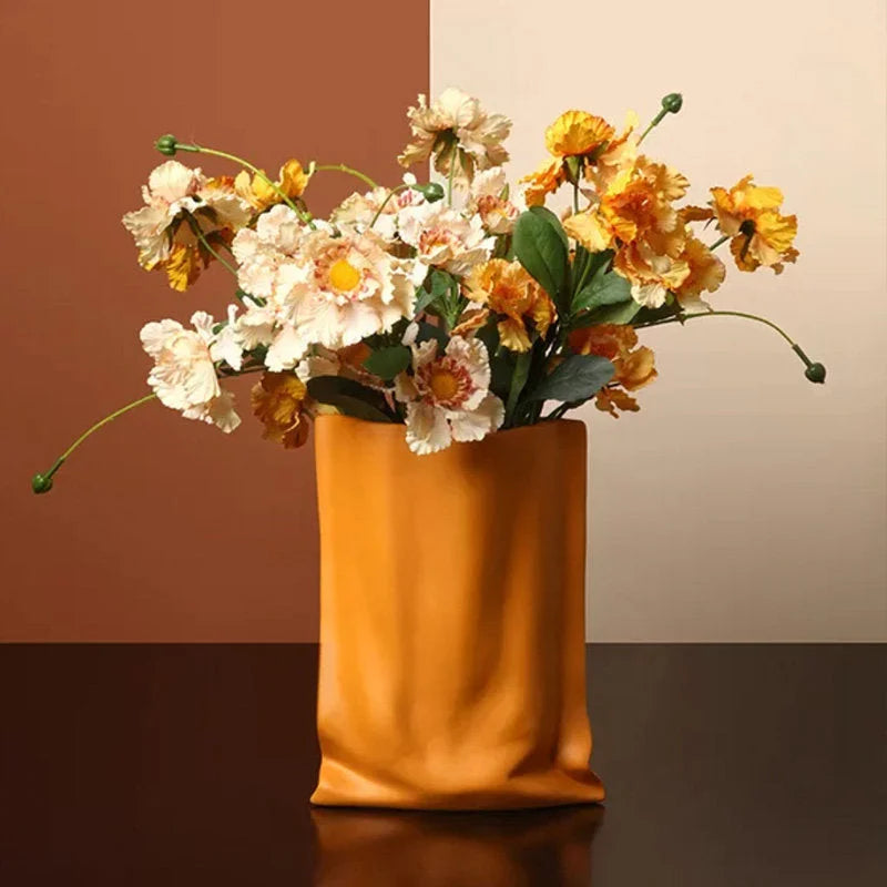 Close-up of the Tangerine ceramic paper bag vase filled with a bouquet of flowers on a dark tabletop against a split-tone background - RoomDen.