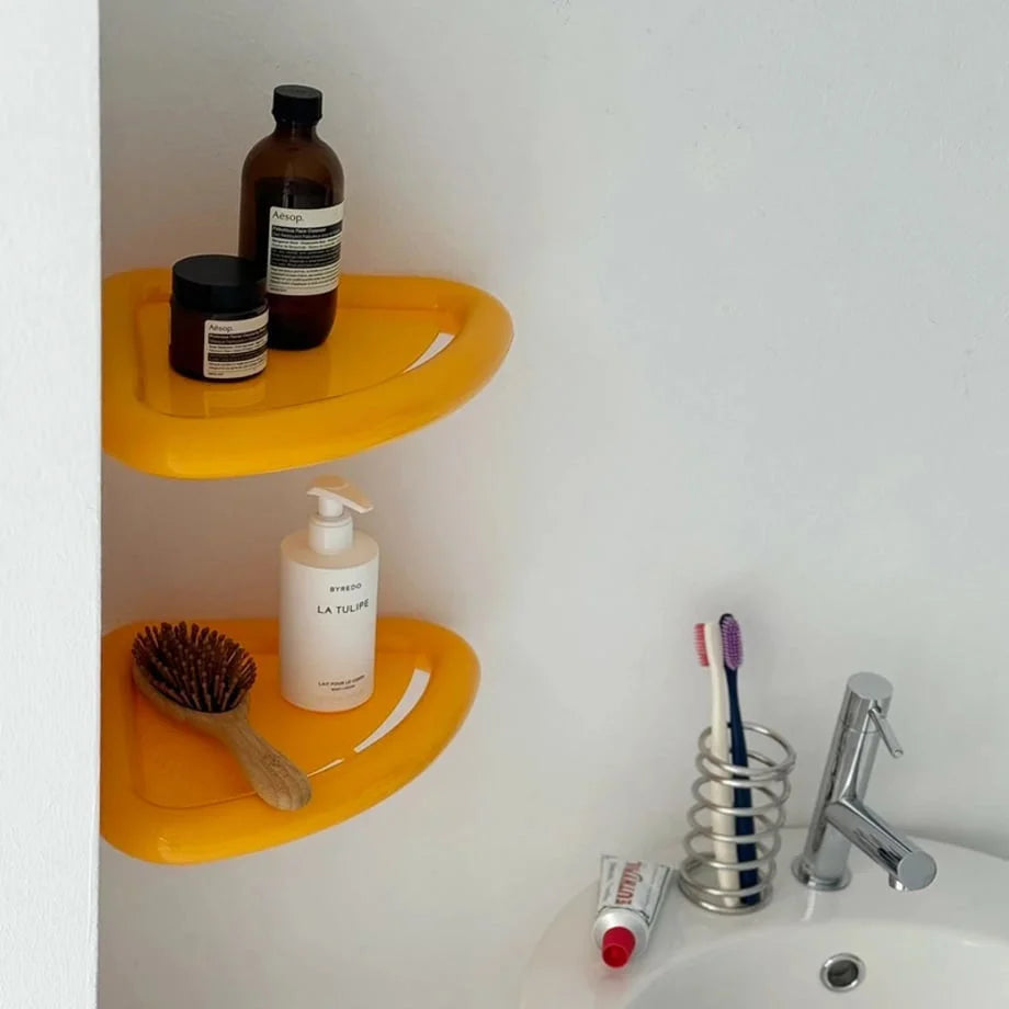 A pair of yellow floating corner shelves displaying skincare bottles and a wooden brush next to a bathroom sink. - RoomDen