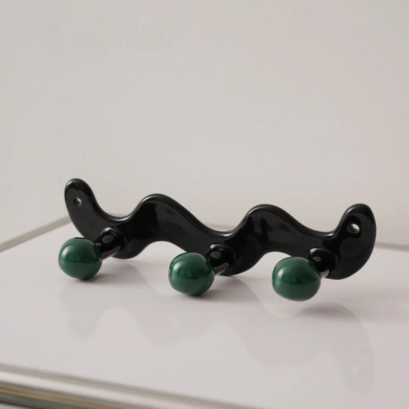 Three-hook wavy black ceramic wall rack with green balls on a white surface - RoomDen.