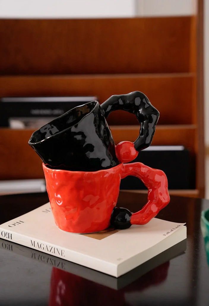 A black and a red textured ceramic mug stacked on a book against a wooden shelf background - RoomDen.