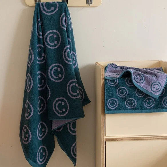 A teal smiley face bath towel hanging from a hook next to a matching face towel folded in a wooden bathroom organizer - RoomDen.