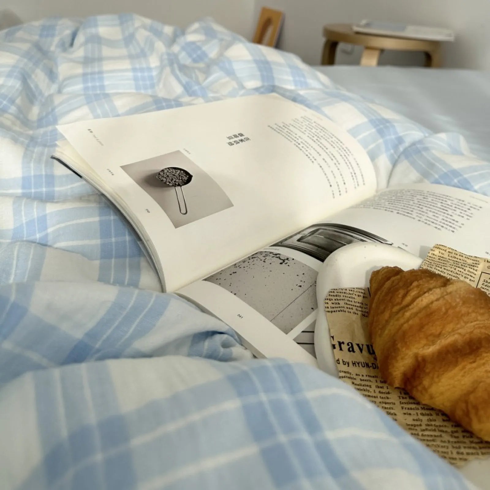 Close-up detail of the soft washed cotton texture of the blue gingham plaid bedding, with a croissant and book nearby.