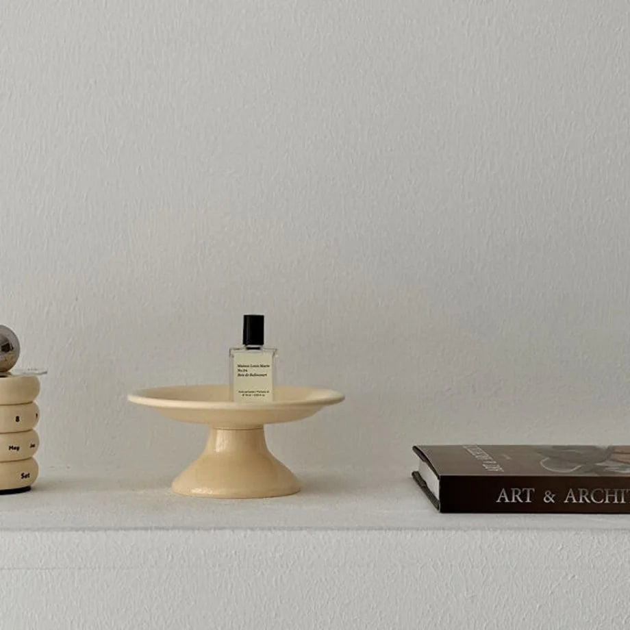 Wider shot of the creamy beige pedestal jewelry dish on a white shelf with a book and calendar. - RoomDen