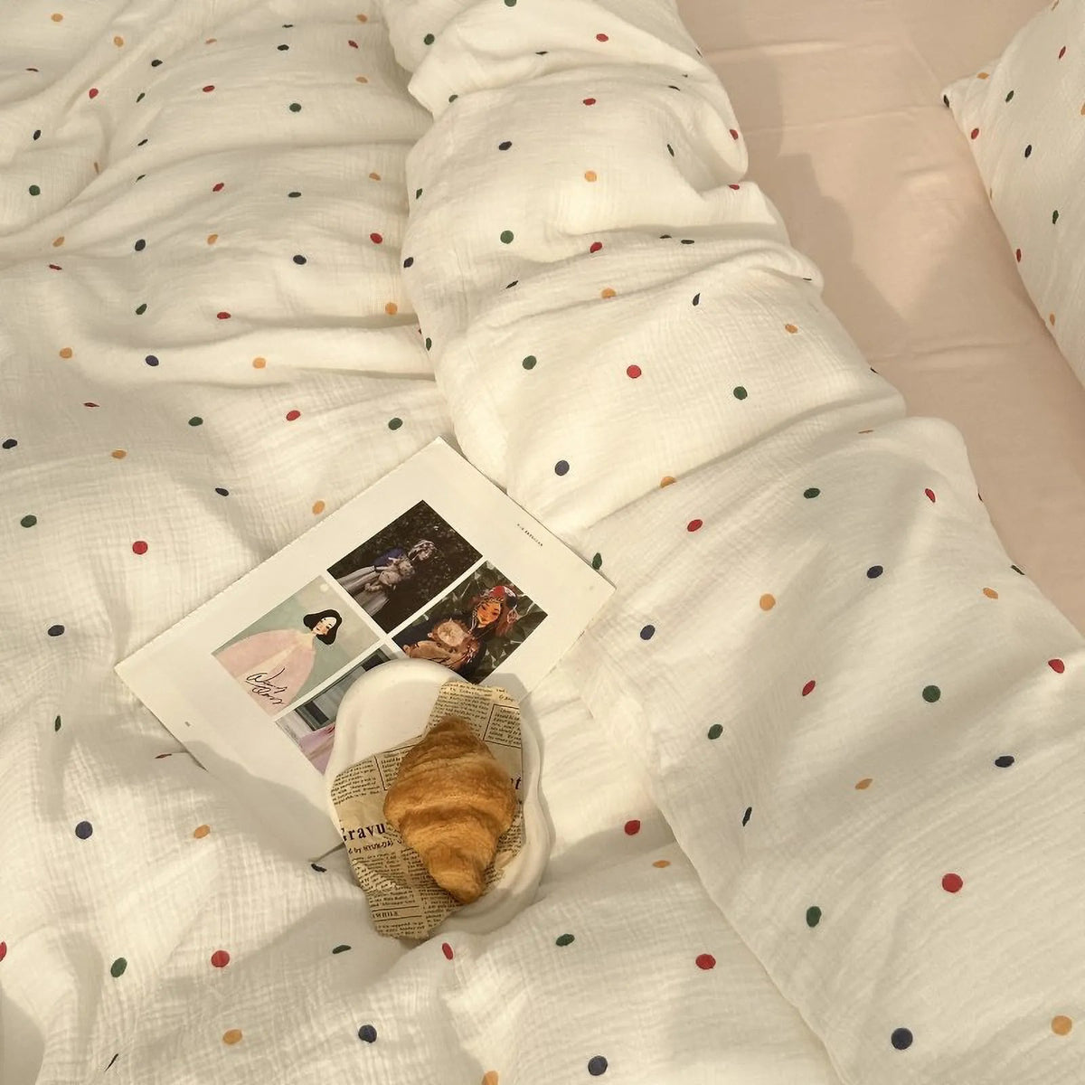 A lifestyle shot of the white polka dot bedding set styled with a magazine and a croissant on a plate. - RoomDen