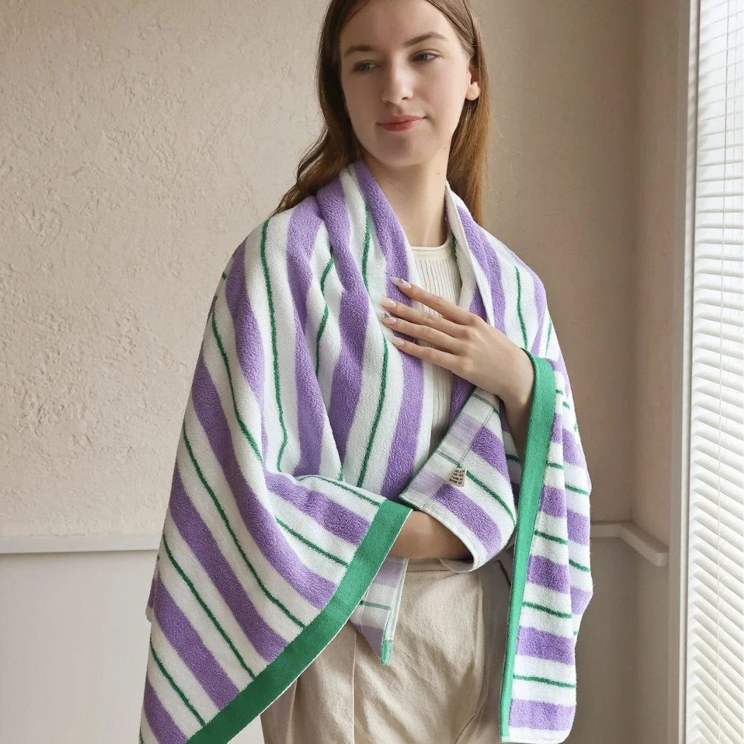 A model wrapping a large purple and green striped cotton bath towel around her shoulders in a brightly lit room - RoomDen.