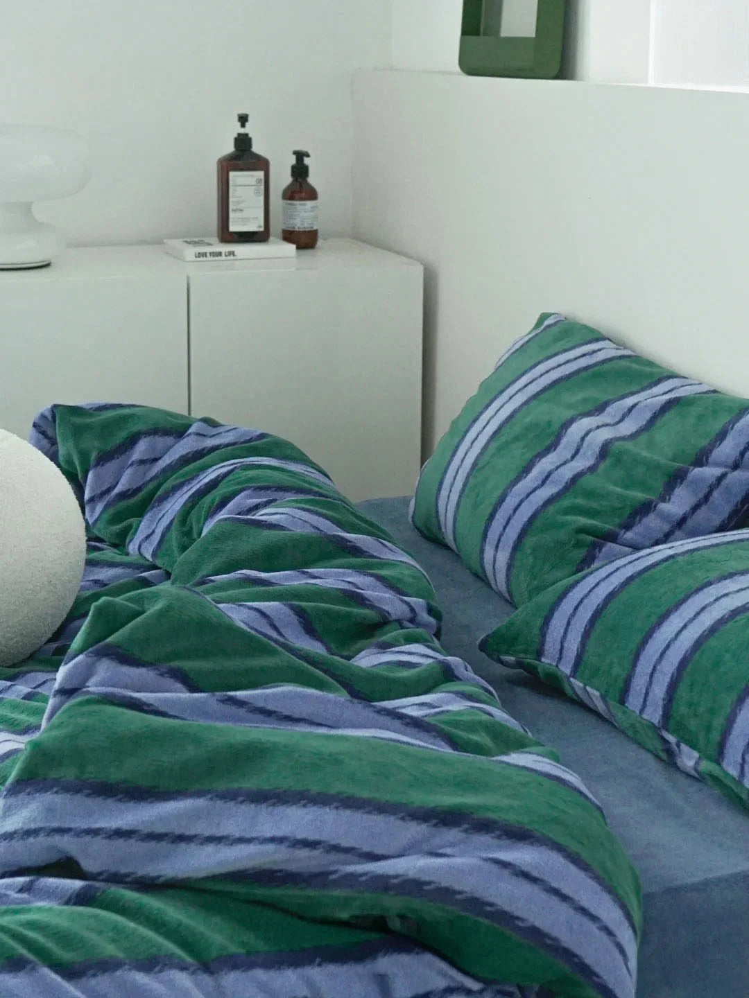 Lifestyle view of the green and blue striped velvet bedding set in a modern white bedroom - RoomDen.