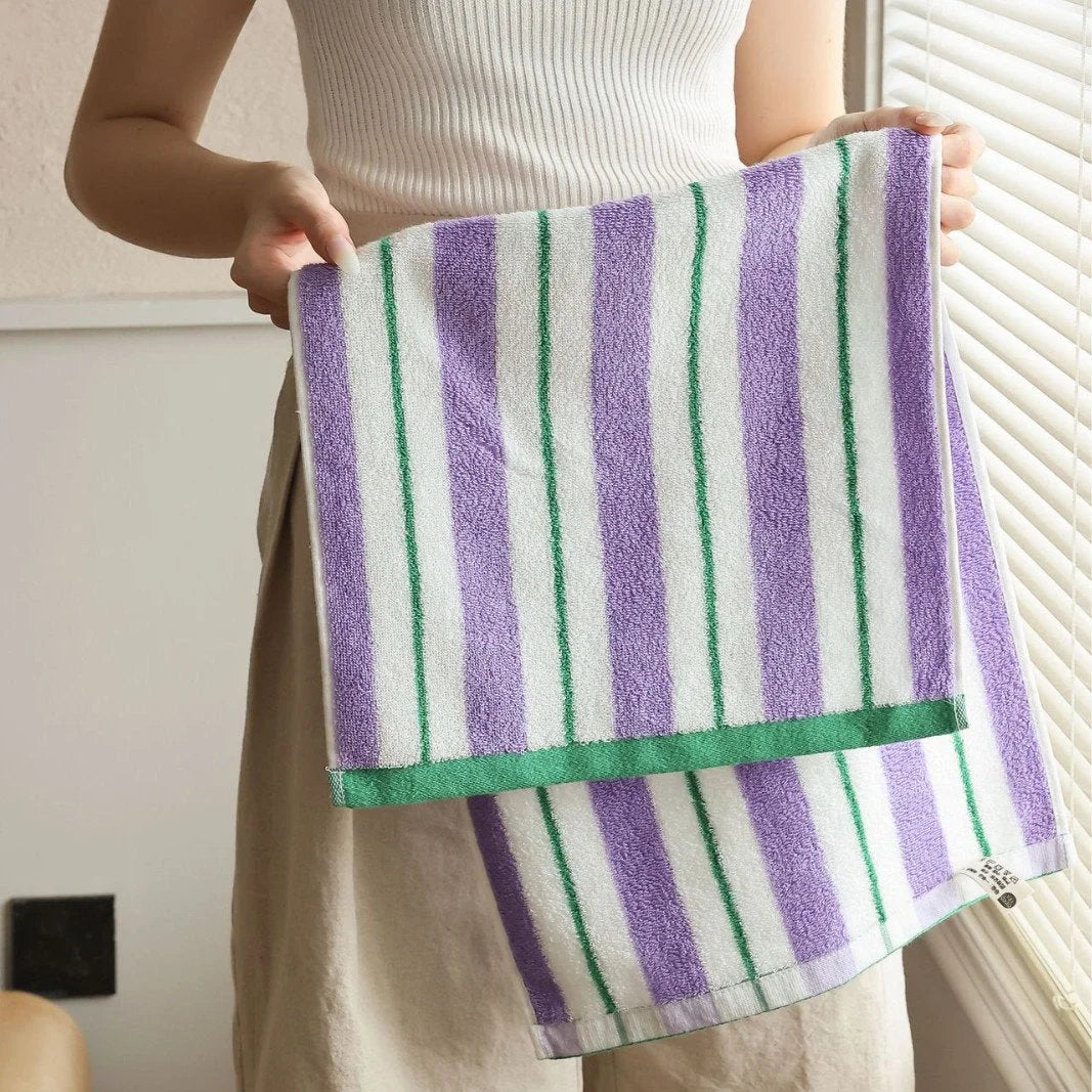 A person holding up a purple, white, and green striped cotton face towel, showing its plush texture - RoomDen.