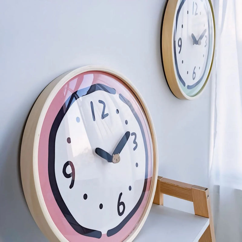 Angled view of the pink and green Whimsical Doodle Style Wall Clocks hanging on a light grey wall - RoomDen.
