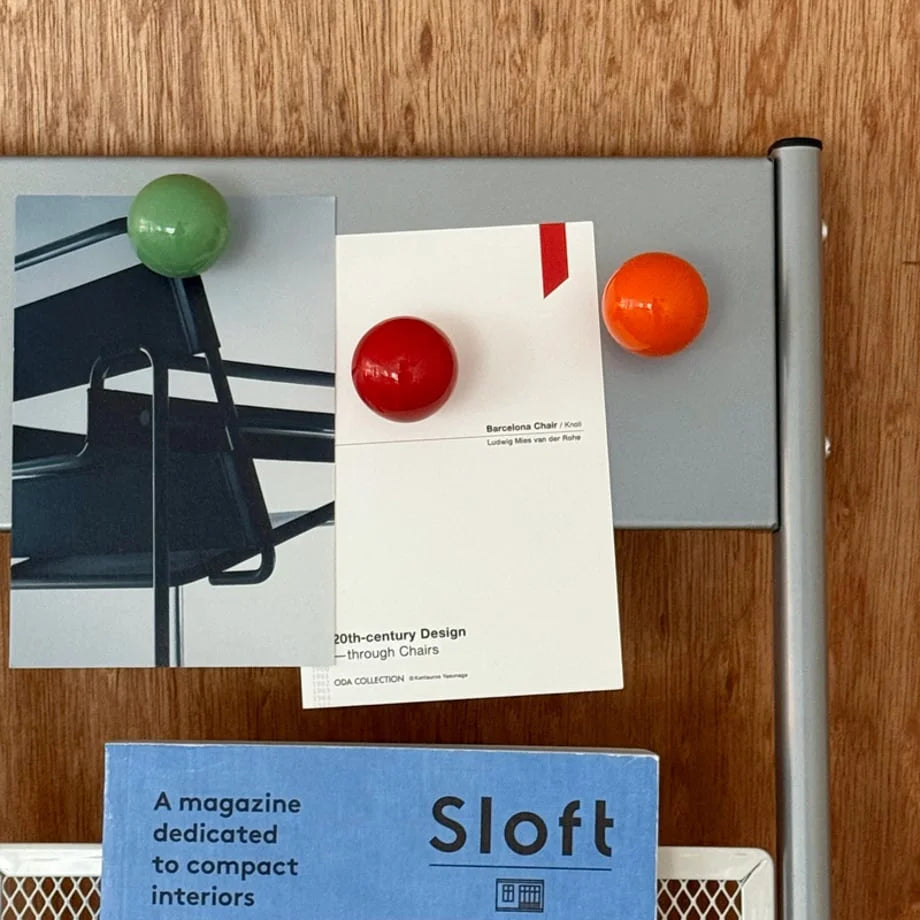 Close-up of Green, Red, and Orange magnetic hooks holding postcards on a grey magazine rack. - RoomDen